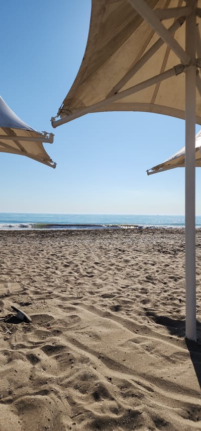 The image depicts a beach scene in Türkmenbaşy, Turkmenistan. The perspective is from under a large, light-colored umbrella, looking out towards the sea. The umbrella's structure, with its white poles and fabric, dominates the left side of the frame. The fabric is a neutral, possibly beige or cream color, and appears to be slightly weathered.

The foreground is the sandy beach, with distinct ripples and patterns formed by the wind or receding waves. The sand is dark and appears to be a mix of fine grains and small pebbles or shells. A single sandal, black and white striped, lies on the sand in the lower right portion of the image.

The middle ground is the calm, light blue sea, meeting the sandy shore with a thin line of gentle waves. The water's surface reflects the bright sky, creating a shimmering effect. The horizon is clear, separating the sea from the pale blue sky above, which suggests a clear and sunny day.

The scene is set outdoors, likely during the daytime, given the bright light and shadows cast on the sand. The overall mood is serene and peaceful, typical of a beach setting. There is no visible text in the image. The focus is on the natural elements of the beach and the man-made structure providing shade.