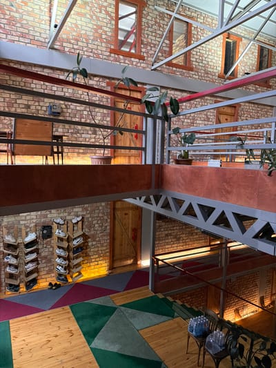 The image depicts the interior of a multi-level building with exposed brick walls and a metal truss ceiling structure. The architecture suggests an industrial or loft-style design. On the lower level, a series of wooden shelves display various pairs of athletic shoes. A patterned carpet, composed of geometric sections of green, burgundy, and grey, is laid out on the wooden floor. In the foreground, a wooden staircase with a metal railing leads upwards, with two chairs visible at the bottom of the stairs, each holding a plastic-wrapped bundle of water bottles. The upper levels feature wooden doors set into the brick walls and metal railings that line the walkways. Potted plants with large leaves are strategically placed along the railings. The lighting appears to be warm, with accent lights illuminating the brickwork. The city of Shabdan, Kyrgyzstan, is mentioned, but there is no visual information in the image that specifically confirms this location.