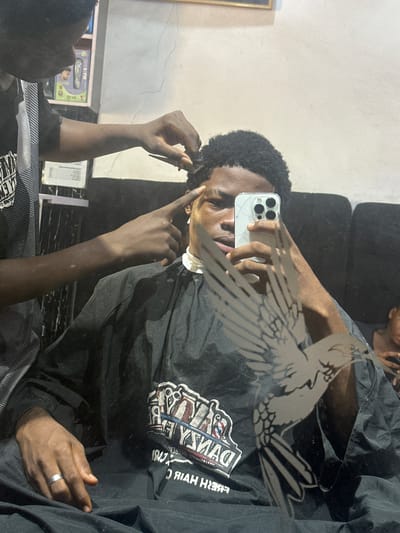 The image captures a mirror selfie taken inside what appears to be a barbershop in Ogwashi-Uku, Nigeria. The main subject is a dark-skinned individual with short, curly black hair, seen from the chest up. They are wearing a black barbershop cape and are holding a white smartphone (likely an iPhone) with a cracked screen protector to take the picture, looking directly into the phone's camera.

The mirror's surface is prominent, featuring a large, stylized graphic of a bird (possibly an eagle or hawk) with outstretched wings, obscuring part of the reflection. The main subject's left hand (as reflected) holds a comb, while another person's hand, presumably the barber or stylist, is seen touching the subject's head, suggesting a haircut or styling in progress. The subject wears a plain silver band ring on their reflected right hand.

The black barber's cape worn by the subject displays a reversed logo that reads "DANKO'S FRESH HAIR" along with a barbershop emblem featuring grooming tools like a razor and comb. In the background, a light-colored wall is visible, along with a partially obscured gold-framed item. Another person is dimly reflected in the lower left corner of the mirror. The setting appears to be an indoor establishment, brightly lit.
