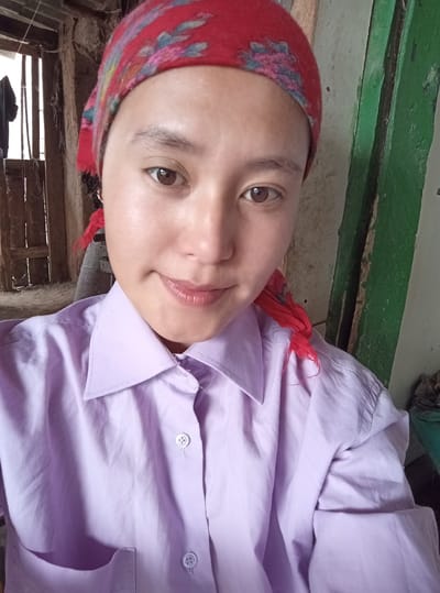 The image is a selfie taken by a person wearing a red patterned headscarf and a light purple collared shirt. The person's face is partially visible, with their eyes looking towards the camera and a slight smile. The background appears to be an indoor environment with rustic wooden structures and green painted elements. There is also a glimpse of another person or figure in the background, further into the room. The overall setting suggests a modest, possibly rural dwelling.