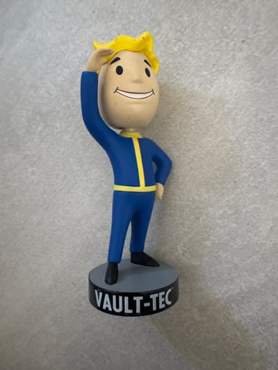This media file features a small figurine of Vault Boy, a mascot character from the Fallout video game series. The figurine is lying down and appears to be on a light gray, textured surface, possibly a floor or tabletop. Vault Boy has bright yellow hair, a cream-colored head with a simple, smiling face featuring black dots for eyes and a curved line for a mouth. He is wearing a blue jumpsuit with yellow trim. The figurine is standing on a circular black base with the text "VAULT-TEC" printed vertically in white letters. The lighting suggests it is daytime, and there are no obvious weather cues. The background is plain and out of focus, with no discernible location details beyond the textured surface. There is no clear activity or event taking place; the figurine is posed statically.
