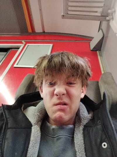A young person with messy brown hair, wearing a gray sweater and a dark jacket with a fleece lining, makes a scrunched face. The individual is seated with a gray headrest behind them. Red walls are in the background, along with a window and a silver vent. A white and silver striped ceiling is visible at the top of the frame. The lighting is bright and appears to be indoor.