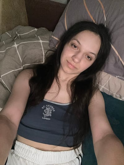 The image shows a young woman lying down on a bed, looking towards the camera. She has long, dark hair and is wearing a navy blue crop top with "ST. GEORGE LONDON" printed on it, and white athletic shorts. She is positioned on her back with pillows behind her head. The background includes a patterned blanket and pillow, and a teal surface. The image does not provide any information that would indicate a specific location within the city of Postoyata, Ukraine.