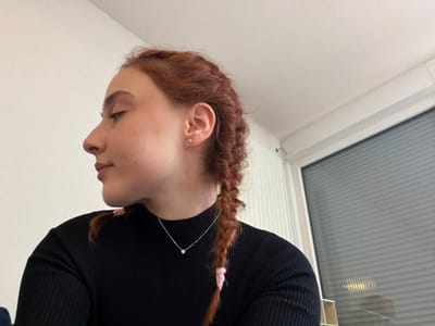 A person is depicted in a partial profile view, facing left, against an indoor setting. The subject is a young individual with reddish-brown hair styled in two braids, parts of which are secured with light pink ties and fall over the right shoulder. They wear a black, ribbed, high-necked long-sleeved top. A thin silver-colored chain necklace with a small, circular, clear pendant is visible at the center of the collarbone. A single, small, clear stud earring is present in the visible right earlobe. The subject's eyes are cast downwards, and the facial expression is neutral to slightly contemplative.
The background consists of a plain white wall occupying the left and upper-left portions of the frame. To the right, a white-framed window with horizontal blinds or textured glass is visible, reflecting an indistinct light source. The ceiling is white. A small metal hook or fixture is affixed to the wall near the top-left corner of the window frame. The specific location within Eschau, France, is not discernible from the visual evidence presented in the image.