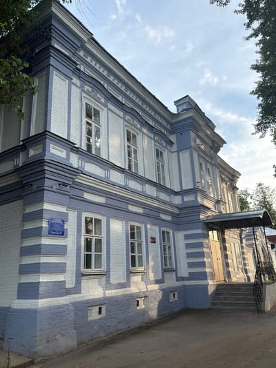 A light blue and white brick building with a dark blue trim is shown under a partly cloudy sky. The building appears to be a historical site, as indicated by a blue plaque on the left side which reads "ПАМЯТНИК АРХИТЕКТУРЫ" (Monument of Architecture) and "Улица Советская, 20" (Sovetskaya Street, 20). This suggests the location is Sovetskaya Street, house number 20, in Cherdyn, Russia. The building has multiple windows, some arched at the top. A covered entrance with a dark metal railing leads to a set of stairs. Trees are visible on the left and right sides of the building, with some branches overhanging the roof. The ground in front of the building is a dirt or gravel surface. There are no people or animals visible in the image. The lighting suggests it is daytime, possibly late afternoon due to the long shadows cast.