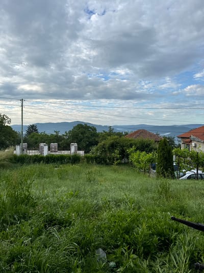The image depicts a rural or semi-rural landscape in Anton, Bulgaria, under an overcast sky with patches of visible blue. The foreground is dominated by unkempt, tall green grass and weeds, with a dark, thin object extending into the frame from the lower right. In the midground, a utility pole stands on the left, supporting multiple overhead wires. Behind a dark fence, a linear arrangement of five light-colored, rectangular stone monuments or markers with dark tops is visible. Dense green trees and bushes surround these structures. To the right, two houses with red-tiled roofs are partially observable; one is largely obscured by foliage, while the other features a white facade and a mounted air conditioning unit. A trellis with green grapevines and a slender evergreen tree stand in front of the latter house, partially concealing a white car. The background reveals multiple layers of hazy, blue-grey mountains stretching across the horizon. The sky is largely covered by varying shades of grey and white clouds, with brighter blue visible in the upper right quadrant. No people are present.