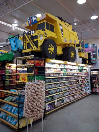 The image depicts the interior of a supermarket in São Gabriel Do Oeste, Brazil. The dominant feature is a large, yellow, decorative dump truck positioned above shelving units filled with various packaged goods. The truck is branded with logos and the number "25". Below the truck, shelves are stocked with food items, including bags of snacks, cereals, and other packaged foods. A prominent yellow sign labeled "OFERTA" (Offer) is displayed on one of the shelves. The store appears well-lit with fluorescent lighting and has a tiled floor. The overall scene suggests a retail environment, possibly with a themed display to attract customers.