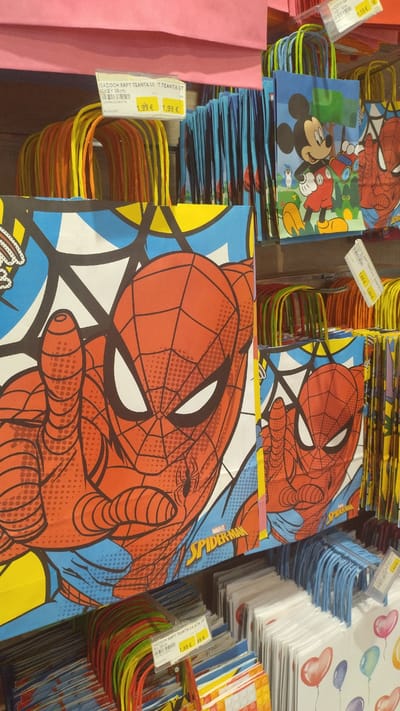 This image shows a retail display of children's gift bags. The bags feature popular cartoon characters such as Spider-Man and Mickey Mouse. Several bags are prominently displayed with their handles showing, indicating they are hanging from a rack. Price tags are visible on some of the bags, indicating a price of 1.99€. The background consists of shelves stocked with more bags and other merchandise, suggesting a store environment. The lighting is typical of indoor retail spaces. The location is Pylaia Municipal Unit, Greece.