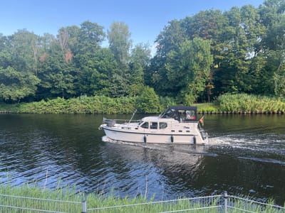 A white motor yacht named "Carolin" travels east on a dark canal, leaving a foamy wake. The boat has a black canopy over the aft deck. The left side of the boat displays the name "Carolin" and the registration "BRB-P59". A German flag is mounted on the stern. The canal is bordered on the north side by a dense line of green trees and bushes, some of which show autumnal brown coloration. On the south side, tall green reeds and grass are visible behind a metal fence in the foreground. The sky is clear and blue. The scene is consistent with the Teltow Canal in Teltow, Germany, a waterway that runs through the Brandenburg and Berlin regions.