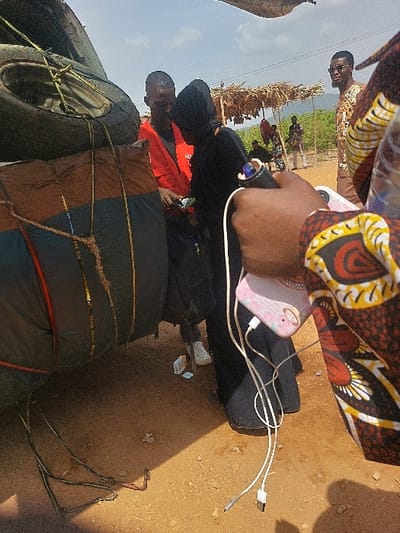The image captures an outdoor scene in Tataura, Nigeria, under a bright, partly cloudy sky. The ground is composed of reddish-brown dirt. In the foreground, positioned to the right, a hand wearing a patterned fabric garment holds a light pink smartphone featuring a cartoon graphic. Two white charging cables are connected to the phone and hang downwards.
To the left, a portion of a heavily loaded vehicle is visible, featuring multiple tires stacked and secured with ropes and bungee cords, along with dark, tarp-wrapped bundles. In the midground, a man wearing a red long-sleeved shirt over a dark shirt, dark pants, and white sneakers stands facing a woman. The woman is dressed in a black abaya and a black hijab, covering her head. They are engaged in an interaction, with the man appearing to show or hand her a small item.
In the background, a rustic shelter with a thatched roof supported by wooden poles is visible. To the right of this shelter, another man, wearing a brown patterned garment and sunglasses, stands and looks towards the camera. Several other indistinct figures are present further in the background, near the shelter and amidst distant green foliage. On the ground, near the woman's feet, a small white box and a blue wrapper are visible. Loose dark cables lie on the ground near the base of the loaded vehicle.