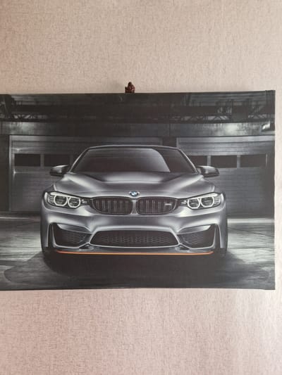 The image shows a poster of a silver BMW M car parked in a garage. The poster is hanging on a textured light-colored wall. A small, red, toy figurine is placed on top of the poster, slightly to the right of the center. There are no discernible features or context that would place this scene specifically in Rikava, Latvia. The image is a close-up of the poster and the wall it is mounted on.