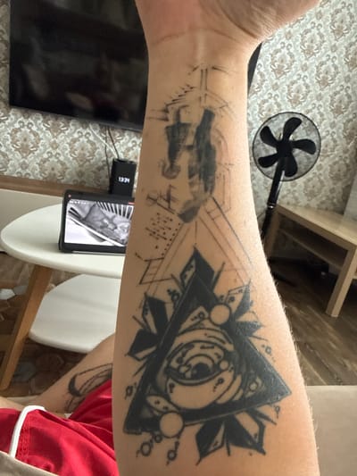 The image displays a person's arm with a tattoo that appears to be a geometric design incorporating an eye within a triangle. There are also other sketched elements on the arm that are less defined, possibly unfinished or a different style of tattoo. The background shows a patterned surface, possibly a wall or fabric, and part of a small, black fan. A shelf unit is visible in the upper right, and a portion of a person wearing a red shirt is in the lower right corner. A tablet or phone screen is visible in the lower center, displaying an image, and a small black device with the text "1334" is attached to it. The setting appears to be indoors, with artificial lighting. No specific time of day or weather conditions can be determined. The emotional tone is neutral, focusing on the visual elements.