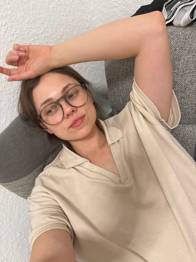 A woman lies on a gray upholstered surface, wearing round eyeglasses and a beige polo shirt. A black and white object is visible in the upper left corner, possibly clothing or footwear. The background consists of a white textured surface, likely a wall. There is no information to indicate the location is in Kőröshegy, Hungary.