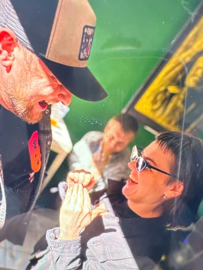 The image displays an indoor scene featuring three individuals, captured with a diagonal perspective. In the foreground on the left, a man is partially visible, wearing a black and tan baseball cap with a dark, indistinct patch. He has a beard and his mouth is open, suggesting he is mid-sentence or reacting with amusement.
To his right, in the mid-ground, a woman is captured in a moment of broad laughter or a cheerful smile, looking downwards and to her left. She wears dark-rimmed glasses and has dark hair with bangs. Her visible left ear has multiple piercings. Her hands are clasped or gestured in front of her, indicating animated conversation. She is dressed in a grey denim-like jacket.
In the blurred background, another person is faintly visible, also appearing to be engaged in the social interaction. The background is dominated by a bright green surface, possibly a wall or screen, exhibiting reflections. Above this green area, a section of a yellow, abstract design or sign is visible. In the lower mid-ground, a distinct red and orange patch, possibly depicting stylized lips or a similar design, is seen on a piece of dark fabric.
The overall atmosphere conveyed is one of joy and lively interaction, suggesting a casual social gathering. The lighting appears to be artificial indoor light, contributing to some glare and reflections visible on the surfaces. No clear, legible text is discernible within the image. The provided context indicates the location is Веревское, Russia, though no specific visual cues within the image confirm this.