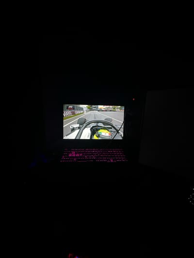 A dark room contains a computer monitor displaying a Formula 1 race replay. A keyboard with pink backlighting is visible to the right of the monitor. The monitor displays a close-up of a Formula 1 car with sponsorship logos, in what appears to be a street circuit. The location of Savigny-en-Véron, France is not represented in the image.