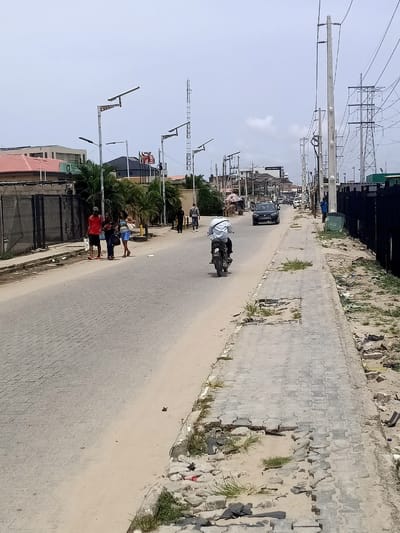 This image depicts a street scene in Eputu Town, Nigeria, under an overcast sky during the daytime. The perspective looks down a paved road, which appears to be made of block pavers, bordered by sandy shoulders.

On the left side of the road, a group of three individuals (two women and one man in a red shirt) walk along a sidewalk. Further down, other pedestrians are visible. In the middle of the road, a person rides a motorcycle away from the viewer, with the word "BOX" visible on its rear. A dark sedan car is seen driving towards the viewer further in the distance.

The right-hand sidewalk is significantly damaged, with many paving blocks missing, exposing sand, sparse grass, and some litter. Utility poles carrying numerous power lines line the right side of the street, leading to large electricity transmission pylons in the background. On the left side, modern street lights equipped with solar panels are installed. Various buildings, including residential and commercial structures, line both sides of the road. In the mid-ground on the left, flags, including what appears to be the Nigerian flag, are visible flying near a building. No other specific text is clearly legible on buildings or signs due to distance and resolution.