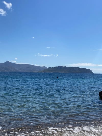This image depicts a coastal scene in Megalochori, Greece, under a bright blue sky with scattered wisps of white clouds. The foreground shows the shallow edge of a dark-colored beach or rocky shore with small white-capped waves gently breaking. The middle ground is occupied by calm, deep blue sea water, rippled by light waves. A single individual's head and upper shoulders are visible above the water in the mid-ground, slightly left of center, indicating a person swimming or wading. To the far right, a dark, jagged rock partially extends into the water. The background presents a panoramic view of distant landmasses across the sea. On the left, a large, dark grey-blue mountainous landmass dominates the horizon, while a distinct, vegetated island or peninsula is visible to the right. A small white sailboat is discernable on the water between the distant landmasses.