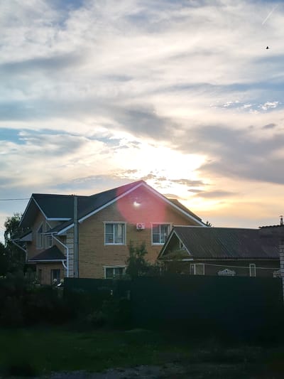 In Selivanovo, Russia, this photograph captures a residential scene with a prominent brick house under a cloudy sky. The sky is filled with a mix of white and gray clouds, with the sun partially visible and casting a warm glow. A single bird is silhouetted against the sky, and a faint contrail from an airplane can be seen.
The main subject is a two-story brick house with a dark roof. It features several windows, some with white trim. An air conditioning unit is mounted on the exterior wall. Adjacent to the brick house, another house with a darker roof and decorative trim is partially visible. Both houses are partially obscured by dark green foliage and a dark green fence in the foreground. The overall impression is that of a quiet, possibly evening, rural or suburban setting.