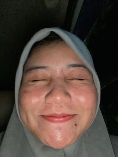 The image is a close-up, selfie-style portrait of a person wearing a light grey hijab, taken in a dimly lit environment. The subject's eyes are closed, and they are smiling. The lighting highlights the contours of their face, revealing some texture and blemishes on the skin. A small dark mole is visible on the right side of their chin. The background is dark and indistinct, suggesting an indoor setting or nighttime outdoors. There is no discernible information about the location within Jawa, Indonesia.