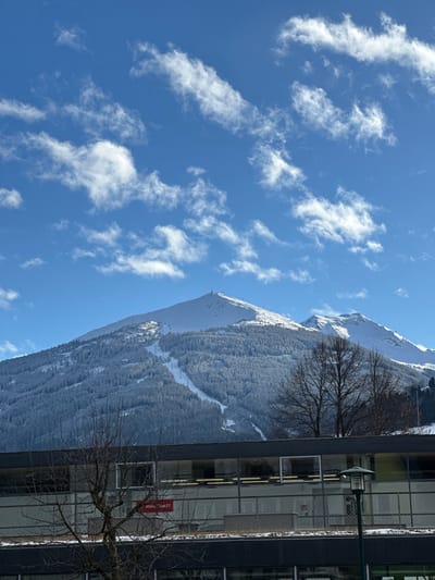This image depicts a winter landscape in Bad Hofgastein, Austria, under a clear blue sky featuring scattered white cirrus and cumulus clouds. A large, snow-covered mountain dominates the background, its slopes covered by dense coniferous forests. A cleared ski piste descends the mountain, with multiple ski lift pylons visible along its path. A structural installation, likely a communication tower, is present at the mountain's peak.
In the mid-ground and foreground, a modern multi-story building with dark exterior fascias and horizontal banding is partially visible. The building features numerous large rectangular windows, some reflecting light, and interior lights are active. A red rectangular sign with white text reading "KNAUS GHIBLET" is affixed to the building's facade. Several bare deciduous trees with intricate branch patterns are positioned in front of the building, partially obscuring it and the lower mountain slopes. An outdoor lamp post with a dark fixture and frosted shade is visible on the right side of the foreground, adjacent to the building. No individuals are present in the scene.
