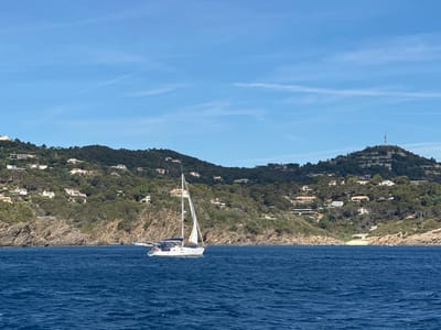 This is an image of the coastline of Bormes-les-Mimosas, France, taken from the water. In the foreground, the deep blue sea has moderate waves. A white sailboat with a single sail is positioned in the middle ground, moving from left to right. The boat has a blue cover over its boom. Behind the boat, a rocky, tree-covered hillside slopes upwards, dotted with numerous white villas. The hillside is densely vegetated with green trees. The sky above is a clear, bright blue with a few wispy white clouds. On the far right of the frame, a distant antenna tower is visible on top of a hill.