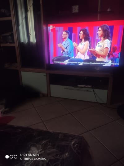 The image shows a television screen displaying a scene with three people. The subjects are positioned vertically, with the top two appearing to be women and the bottom one a man. The woman at the top is wearing glasses and a white t-shirt, with dark curly hair. The woman below her also has dark curly hair and is wearing a light-colored dress. The man at the bottom is wearing glasses and a blue shirt. The background on the TV screen appears to be a colorful, stylized set with horizontal pink and purple bands. The TV itself is housed in a wooden media stand, and the room's tiled floor is visible in the foreground. A portion of a dark, patterned fabric or blanket is also in the foreground, partially obscuring the tiled floor. The lighting suggests it is likely indoors and evening, as the TV screen is illuminated and the rest of the room appears dimly lit. Text visible on the screen in Italian mentions "realizzato dalla direzione," which translates to "produced by the direction." Additionally, a watermark in the bottom right corner reads "SHOT ON MI 9T AI TRIPLE CAMERA."
