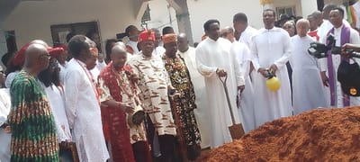 This image captures an outdoor ceremonial gathering in Umu Enge, Nigeria, under bright daylight. A diverse group of people, primarily men, are present, suggesting a formal event.
In the foreground, a prominent mound of reddish-brown earth indicates an activity related to ground-breaking, a foundation ceremony, or possibly an interment. Several individuals are dressed in traditional Nigerian attire, featuring colorful patterned fabrics and distinctive headwear, such as red beaded caps or crowns, with some holding ceremonial staffs.
A significant number of men are dressed in white robes, some of whom appear to be clergy, with one on the far right wearing a purple stole over his white vestment. One man in white robes is seen holding a small, metal shovel with its tip resting in the earth pile, signifying an act of digging or turning soil. Another man in white holds a bright yellow object, possibly a small container or sprayer, which might be used for ritualistic sprinkling.
A building is visible in the background. The overall scene conveys a sense of solemnity and attention, typical of a community or religious ceremony. On the far right, a portion of camera or video recording equipment is visible, suggesting the event is being documented. No text is discernible within the image.