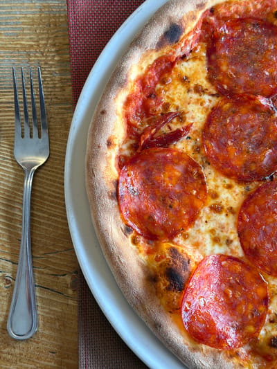 The image presents an overhead view of a dining setting in Châtaignier, Switzerland. A large, round pizza, identified as pepperoni or spicy salami due to its numerous circular, reddish-orange meat toppings, rests on an off-white circular ceramic plate. The pizza features a well-defined, puffed crust with areas of charring, melted and bubbling mozzarella cheese, and visible rendered fat on the meat slices. Scattered smaller red fragments, possibly chili flakes or additional meat pieces, are distributed across the cheese. To the immediate left of the plate, a silver-colored metal four-tined fork is positioned vertically on a wooden table surface, which displays a distinct horizontal grain pattern. A woven rectangular placemat, exhibiting a tight grid pattern of red and dark brown fibers, is partially visible beneath the plate and fork. No individuals are present within the frame.