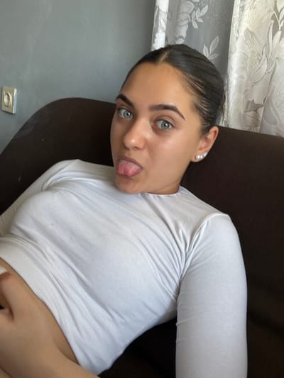 The image shows a close-up, chest-up view of a young woman with dark hair pulled back, blue-green eyes, and fair skin. She is wearing a white, long-sleeved shirt and has a pearl earring in her visible left ear. Her tongue is sticking out. She is positioned on a dark brown couch, with a grey wall and a lace curtain visible in the background. There is a light switch plate on the grey wall. The scene appears to be indoors, likely in a residential setting. There is no indication of the specific location within the city of Radievo, Bulgaria.