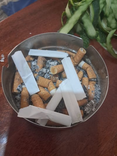 An ashtray full of cigarette butts and ash sits on a wooden table. Above and behind the ashtray, a pile of green vegetable peels is visible. The scene appears to be indoors, possibly in a home or restaurant setting in Chobruchi, Moldova. There are no people or active interactions depicted in the image.