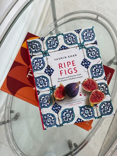 The image depicts a stack of books on a glass-topped table. The uppermost book, titled "Ripe Figs" by Yasmin Khan, features an image of figs on its cover. Underneath this book, another book can be partially seen, with a maroon and orange color scheme. The surrounding environment appears to be an indoor setting, given the nature of the furniture. Based on the location specified, the scene is likely within the city of Ghadeer Al Tayr, United Arab Emirates, but nothing in the image confirms this.