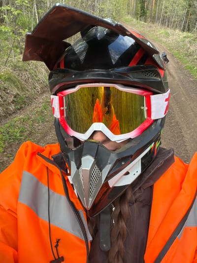 A person wearing a black, red, and white motocross helmet with reflective gold goggles is positioned outdoors. They also wear an orange high-visibility jacket with reflective stripes. A dark braid of hair is visible emerging from beneath the helmet. The surrounding environment includes a dirt path or road, trees, and green vegetation. The location is in a wooded area, with the reflected image in the goggles showing a similar landscape of trees and foliage. Given the presence of trees and dirt roads, the scene likely takes place in a natural or semi-natural setting within Manskii, Russia.