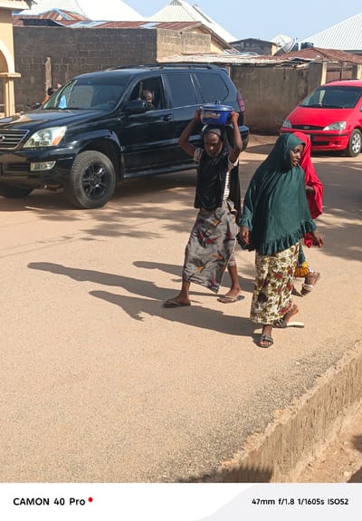 The image shows three people walking on a paved street in Wase, Nigeria. A young person in the center carries a blue pot on their head, wearing a black top with white stripes and a patterned skirt. Another person to the right wears a green headscarf and a patterned skirt. Part of a third person wearing red is visible as well. A black SUV is parked to the left, and a red car is parked behind them. The background shows various buildings with metal roofs and brick walls. The scene is brightly lit with sunlight, casting shadows of the people on the street.