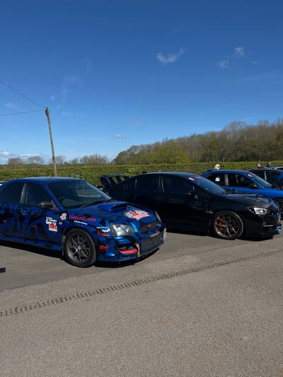 The image depicts several cars parked on an asphalt surface, likely a road or parking area, under a clear blue sky with sparse clouds. The cars appear to be rally or racing-modified vehicles. The foremost car is predominantly blue with a camouflage pattern, adorned with numerous decals and stickers. Behind it is a black car, followed by another blue vehicle, both oriented in the same direction. Tire tracks are visible on the asphalt, running along the length of the parked cars. A row of trees lines the horizon in the background. The image does not offer specific clues to pinpoint the exact location within Silverstone, United Kingdom. No people are clearly visible. The scene suggests an event or gathering related to motorsports or racing.