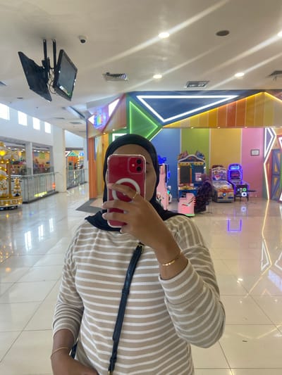 A person wearing a black hijab and a striped long-sleeved shirt is taking a selfie in a brightly lit indoor amusement arcade, likely within a mall in Kudus, Indonesia. The person is holding a red smartphone in their right hand. In the background, various arcade machines are visible, with colorful walls and neon lighting decorating the space. To the left, a carousel or similar amusement ride can be partially seen. The floor is highly reflective, mirroring the lights and the arcade's decor. Overhead, several monitors and a security camera are mounted on the ceiling.