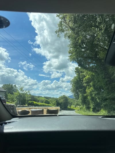 The image shows a view through a car windshield on a sunny day in Westport, Ireland. In the foreground, the dashboard of the car is visible, with a "Mercedes-Benz" sticker on the windshield. Beyond the windshield, a rural landscape unfolds. A stone wall runs along the side of the road, with a car parked behind it. Lush green trees and rolling hills dominate the mid-ground and background. The sky is a vibrant blue, dotted with fluffy white clouds. Power lines are visible stretching across the sky. A distant mountain peak can be seen in the far background. The overall impression is one of a peaceful, scenic drive through the Irish countryside.