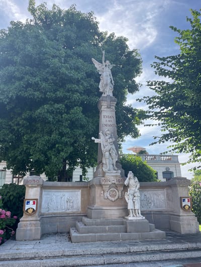 The image shows a war memorial located in Évian-les-Bains, France. The memorial is a tall, stone monument topped with a statue of an angel. Inscriptions on the monument include "EVIAN À SES ENFANTS" and dates "1914-1918" and "1945". There are also relief sculptures depicting scenes of war and figures of soldiers and civilians. The memorial is surrounded by trees and a building with a balcony. The sky is blue with some clouds. The overall scene depicts a solemn tribute to those who served and died in conflicts.