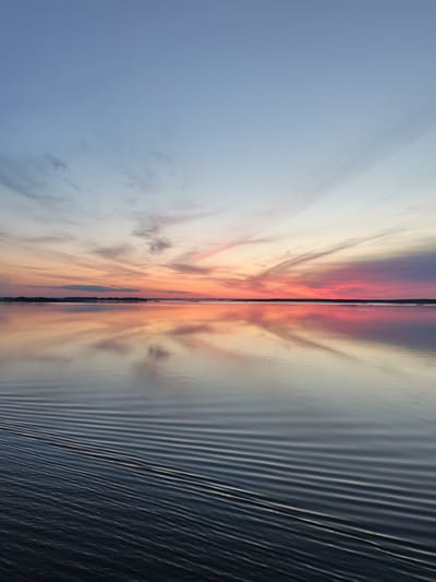 This is a vertical, low-angle, full-shot photograph of a lake at sunset, near Chaika, Russia. The sky displays a gradient of colors, transitioning from a pale blue at the top to vibrant pinks and oranges near the horizon. Wispy clouds are scattered across the sky, catching the sunset's light. The calm water of the lake perfectly reflects the colorful sky, creating a mirror image. In the foreground, the dark water is disturbed by gentle ripples, possibly from a boat. A thin, dark line of trees or land is visible along the horizon. The overall scene is serene and peaceful.