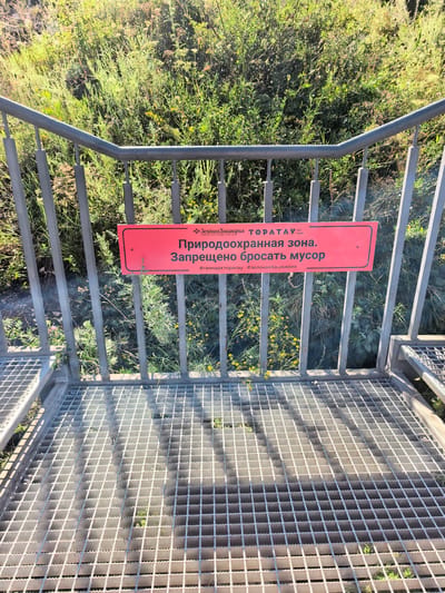 The image shows a sign attached to a metal railing. The sign is red and rectangular with white text in Russian. The text reads: "Зелёная Башкирия ТОРАТАУ ГЕОПАРК Природоохранная зона. Запрещено бросать мусор #геопаркторатау #зеленаябашкирия". This translates to: "Green Bashkiria TORATAYU GEO PARK Protected zone. Littering is forbidden #geoparktoratau #greenbashkiria". The railing is part of a walkway or viewing platform with a metal grate surface. In the background, there is lush green vegetation with some yellow flowers. The environment appears to be outdoors, possibly in a natural park or protected area. The city of Akhmerovo, Russia, is mentioned in the context of this location.