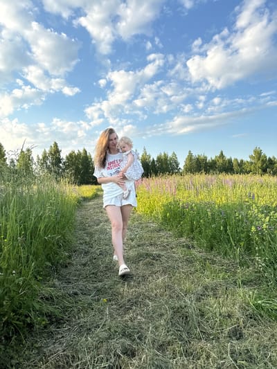 A woman with long, wavy blonde hair is holding a blonde toddler in her arms as they walk along a mown grass path. The woman is wearing a white t-shirt with red lettering that reads "APRES INSTINCT" and white shorts. The toddler is wearing a white and blue patterned dress. They are in a field with tall green grass and purple and yellow wildflowers. In the background, there is a line of trees under a blue sky with scattered white clouds. The scene appears to be in a rural outdoor setting.