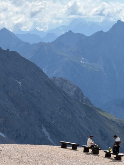 This is a photograph taken from a high vantage point in the municipality of Wildermieming, Austria, overlooking a vast mountain range. The foreground consists of a gravelly, rocky surface, suggestive of a mountain path or viewpoint. Three dark, wooden benches are arranged on this surface. Two individuals are present. On the middle bench, a man with dark hair and a beard, wearing a light-colored t-shirt, is seated, looking towards the camera. He has a green bag beside him. On the rightmost bench, another man, wearing a white t-shirt with dark stripes and black pants, is standing, interacting with a black backpack and some bottles. The middle ground is dominated by the steep, rocky slope of a mountain, showing varied textures and some patches of snow. The background features a layered panorama of receding mountain peaks, fading into a hazy blue atmosphere, indicative of atmospheric perspective. The sky above is partially cloudy.