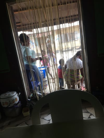 The image depicts a street scene in Awjaw, Nigeria, viewed from a dimly lit interior through a doorway or large window partially covered by a beige string curtain.
In the midground, from left to right, are three individuals. A woman stands on the left, wearing a light blue t-shirt and blue jeans, with her hair pulled back. She faces right, looking towards the exterior. Near her, a blue bucket is visible on the ground. A young child, dressed in a red long-sleeved shirt and dark pants, stands facing away from the camera, looking towards the street. To the right, another woman is seated on a wooden stool, facing away from the camera. She wears a white t-shirt and a multi-colored striped skirt (green, purple, white) and has dark hair, possibly dreadlocks or braids.
In the background, beyond the immediate figures, an unpaved street or pathway is visible. Several indistinct figures can be seen further down the street, including what appears to be another child in a pink top. Buildings with corrugated metal roofs are visible across the street under daylight conditions.
In the left foreground of the interior, a white enamel pot with colorful decorative patterns and a lid rests on a larger purple or grey plastic container. A small yellow container is on the floor nearby. The immediate foreground is partially obscured by the back of a light-colored, possibly white, plastic chair. The interior floor features patterned tiles.