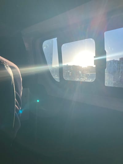 The image depicts the interior of a vehicle, likely a train, bus, or large van, with the sun low in the sky and directly visible through the windows, creating significant lens flare. The perspective is from within the vehicle, looking out.
On the left side of the frame, a portion of a person's upper body, specifically a shoulder and arm in light-colored clothing, is visible in silhouette. The rest of the vehicle's interior is largely underexposed and dark. A prominent blue-green lens flare appears in the lower-left quadrant.
Through three distinct, vertically oriented windows with rounded corners, a hazy exterior scene in the city of Monastir, Tunisia, is visible. The bright, overexposed sun is centered in the middle window, obscuring direct detail. Distant buildings, including what appears to be a multi-story structure on the far right, are partially discernible against the bright sky. The windows themselves exhibit smudges or dirt. No specific actions or interactions are captured.