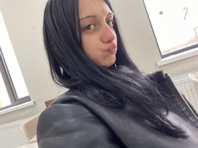 A female subject with long, dark hair, dark eyebrows, and brown eyes is depicted in a medium-close-up shot, looking directly at the camera with a pouting expression. She is wearing a black leather-like jacket featuring a silver zipper and a dark fur-like collar lining. Her hair falls over her left shoulder and chest. The background shows an indoor environment with light-colored walls. Two windows are visible, admitting natural light. The right window pane displays two festive decorations: a red and white striped candy cane shape and a smaller circular red and white striped object. A white radiator is positioned beneath the right window. A light-colored upholstered chair is partially visible on the left side of the frame. The scene is located in ლესის თემი, Georgia.