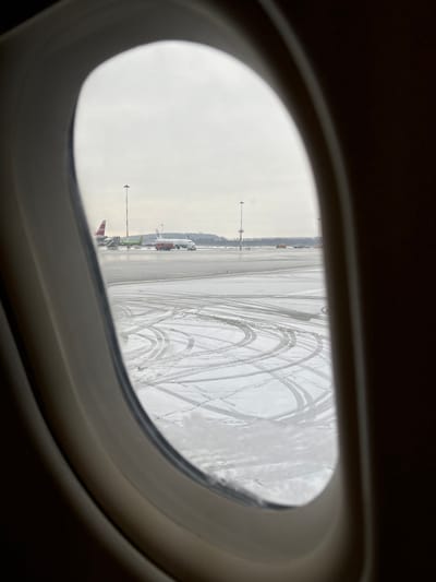 The scene depicts an airport apron viewed from inside an aircraft through an oval window, likely in Novogorelovo, Russia. The external environment is overcast with a muted sky, characteristic of a winter day. The ground outside is partially covered in snow, exhibiting numerous dark tire tracks from ground vehicles, particularly in the foreground and midground areas. Patches of dark, wet asphalt are also visible.

In the midground, two commercial passenger aircraft are present. To the left, a white aircraft with a prominent red tail fin and a green logo (S7 Airlines livery) is stationary, connected to a jet bridge. Ground support equipment, including a set of airstairs and other vehicles, are positioned near this aircraft. To the right of the S7 aircraft, a second white aircraft with a blue and red tail logo (likely Rossiya Airlines, part of Aeroflot Group) is on the tarmac. A red ground service vehicle, possibly a tug or de-icing truck, is positioned near the nose of this aircraft.

Multiple tall, dark grey light poles with arrays of luminaires stand across the apron. In the far background, a dark line of trees or forest is visible under the hazy sky, marking the perimeter of the airfield. No individual people are distinctly identifiable, but their presence is indicated by the operational aircraft and ground vehicles. The overall environment suggests an active airport during cold weather conditions.
