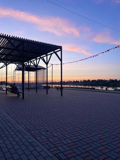 The image depicts a waterfront promenade in Sovetsk, Russia, at sunset. The sky is a gradient of purple and pink, with wispy clouds. A dark, modern gazebo with a slatted roof stands to the left, partially covering two wooden benches where two people are seated, facing away from the camera. The ground is paved with red and dark gray bricks laid in a herringbone pattern. In the background, a river is visible, with a treeline on the opposite bank. A low metal fence runs along the riverbank. Small, triangular flags strung on a wire stretch across the upper right portion of the sky. A stage structure with a white screen is set up behind the gazebo. The scene is tranquil, with the warm colors of the sunset reflecting on the water and creating a peaceful atmosphere.