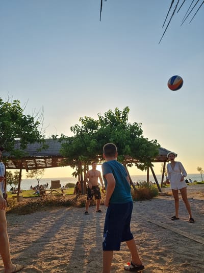 In Palmachim, Israel, at a beachside location with a sandy ground, four people are playing volleyball. The sun is setting, casting long shadows across the sand. A volleyball is in mid-air, about to be hit. In the foreground, two young males are visible, one from the back wearing a blue t-shirt and dark shorts, and another partially visible on the left. In the background, a shirtless male wearing patterned shorts stands to the right, and a female in a white jumpsuit and a flower crown is on the far right. Behind them, a covered structure with wooden beams and a thatched roof provides shade, and further in the distance, people are seated and walking near the ocean. The sky is clear and a pale blue, with a sliver of the sun visible near the horizon.