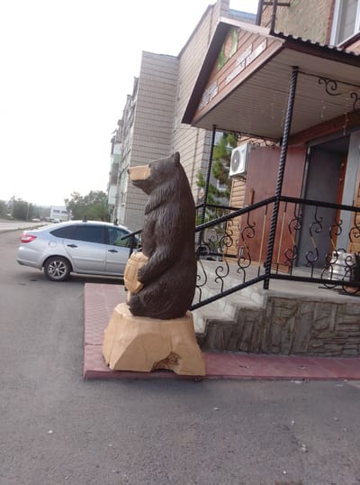The image shows a wooden carving of a bear holding a barrel in front of a building in Shemonaikha, Kazakhstan. The bear carving is positioned on a wooden base, which is placed on a red tiled platform. Behind the carving, a silver sedan car is parked on the asphalt road. The building appears to be a commercial establishment, with a porch featuring a metal railing and steps leading up to an entrance. An outdoor air conditioning unit is visible on the side of the building. The sky is overcast, and trees are visible in the background. The overall scene depicts an outdoor urban environment.