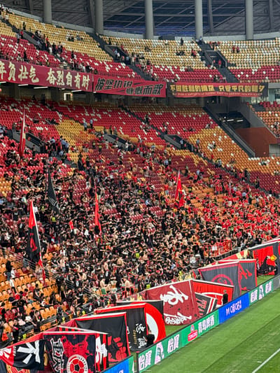 The image shows a large crowd filling the stands of a stadium with banners. The seating area is filled in with a complex pattern of red, gold, and orange colors. Many people wear black attire. Some banners contain Chinese writing and English translations. One banner says "NOT BEING A SUPPORTER OF VICTORY, ONLY LOVE ORDINARY DAYS AND NIGHTS." Other banners depict sports imagery and the word "Loyalty." The image's focus is on the densely packed crowd and the visual impact of the banners and seating arrangement, suggesting an event or gathering in Longxing, China.