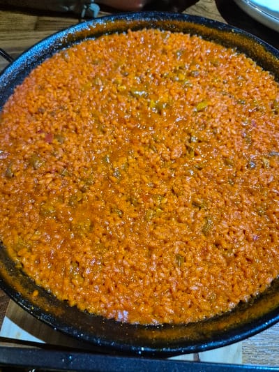 A circular, dark-colored pan, characteristic of a paella pan, is centrally positioned and filled with an orange-red rice dish, likely located in Piedrahíta, Spain. The rice grains are uniformly cooked and coated in a vibrant, viscous sauce. Small, indistinct, darker fragments, possibly vegetables or other ingredients, are visible throughout the rice. The surface of the dish exhibits a moist, reflective sheen. The pan rests on a light-colored, possibly wooden, table surface. The background is out of focus, revealing blurred elements, including a light-colored object in the upper right quadrant and a dark, indistinct bar along the bottom edge of the frame. No individuals are present, and no specific actions are depicted.
